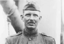 A man in a military dress uniform stands aboard a ship.